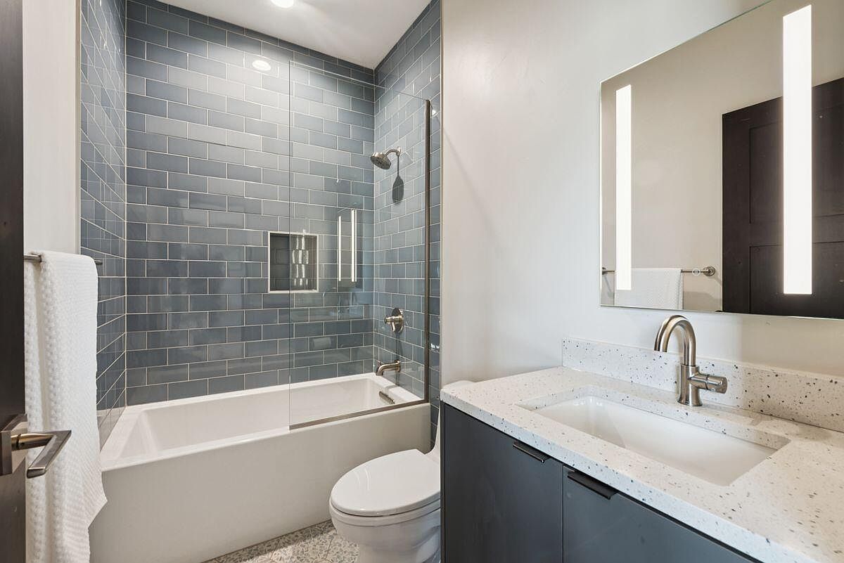 This is a well-lit bathroom featuring a bathtub with blue subway tile surround and a glass partition. A modern vanity with a speckled countertop and gray cabinets is visible, along with a sleek mirror with integrated lighting. The overall design is contemporary and clean, creating a relaxing and functional space.