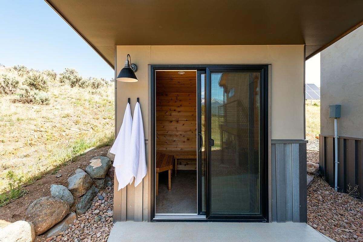 This image showcases a modern patio area featuring a sauna accessible through a sliding glass door. The exterior is finished with a combination of light beige siding and gray vertical paneling. A black barn-style light fixture illuminates the entrance, and two white towels hang neatly beside the door, adding a touch of comfort and convenience.