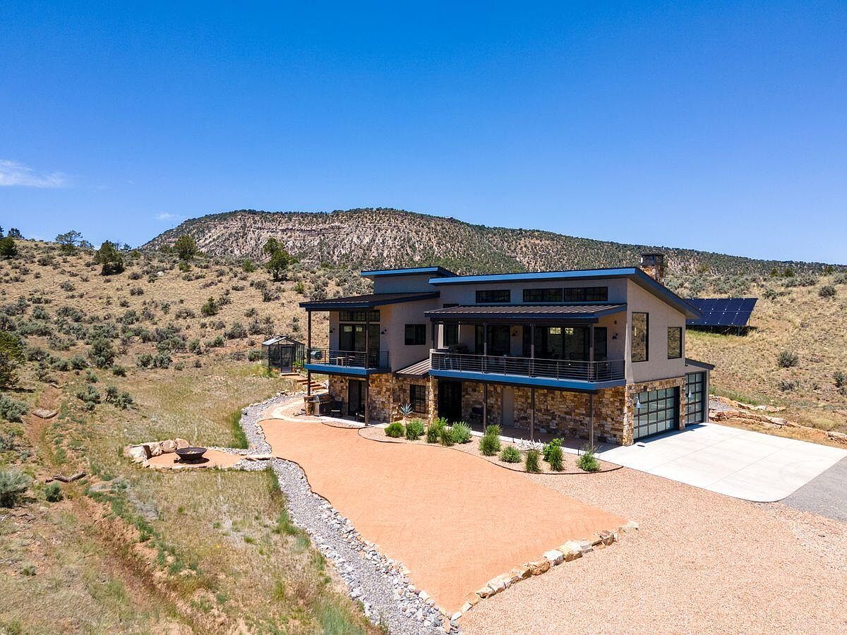 This aerial view showcases a modern two-story home nestled in a natural landscape. The property features a combination of stone and siding exterior, a spacious driveway, and a large outdoor area with a fire pit. Solar panels are visible on the hillside, highlighting the home's energy efficiency.