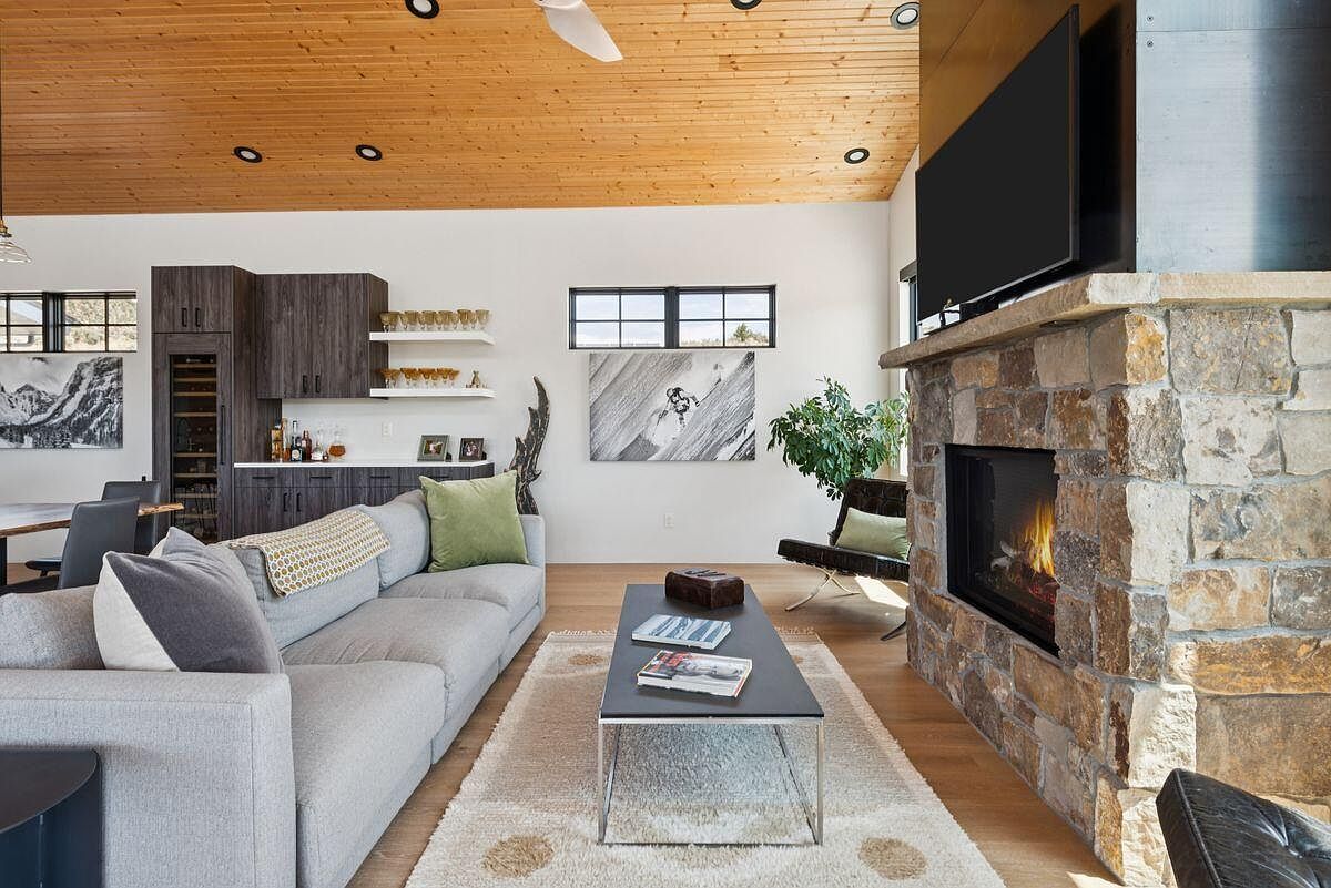 This is an interior shot of a living room featuring a stone fireplace with a mounted television above it. The room is furnished with a gray sectional sofa, a modern coffee table, and a leather chair, all set on a light-colored rug. The ceiling is wood paneled with recessed lighting, and there is a built-in bar area with wine storage, creating a cozy and inviting atmosphere.