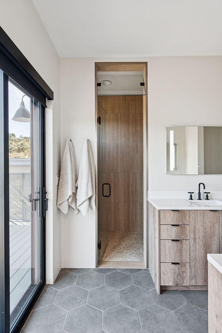 This is a modern primary bathroom featuring a wood-look vanity with a white countertop and a black faucet. A frameless glass shower with vertical wood-look tiling is visible, and gray hexagonal floor tiles add a contemporary touch. A sliding glass door leads to an outdoor deck, enhancing the spa-like atmosphere.