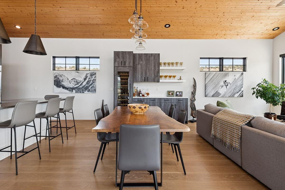 This is an interior shot of a dining room featuring a large, natural-edge wooden table surrounded by modern chairs with black legs. The room has a contemporary design with a wooden plank ceiling, white walls, and hardwood floors. A built-in bar area with wine storage and artwork adds to the sophisticated ambiance, while a glimpse of the living room with a sofa and indoor plant suggests an open-concept layout.