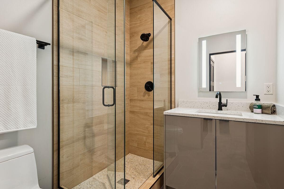 This is a modern primary bathroom featuring a glass-enclosed shower with wood-look tile and a speckled stone floor. The vanity has a light countertop and taupe cabinets, complemented by a sleek, illuminated mirror. The overall design is clean and contemporary, creating a spa-like atmosphere.