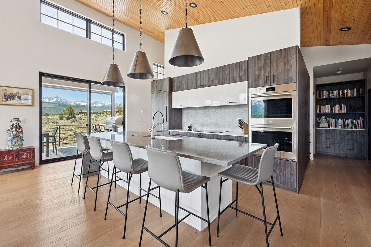 This is a bright and modern kitchen featuring a large island with bar seating, sleek cabinetry in a dark wood grain and white, and stainless steel appliances. The open floor plan allows for views of the outdoor scenery through a large sliding glass door, and pendant lighting adds a touch of elegance. The hardwood floors and wood-paneled ceiling contribute to a warm and inviting atmosphere.