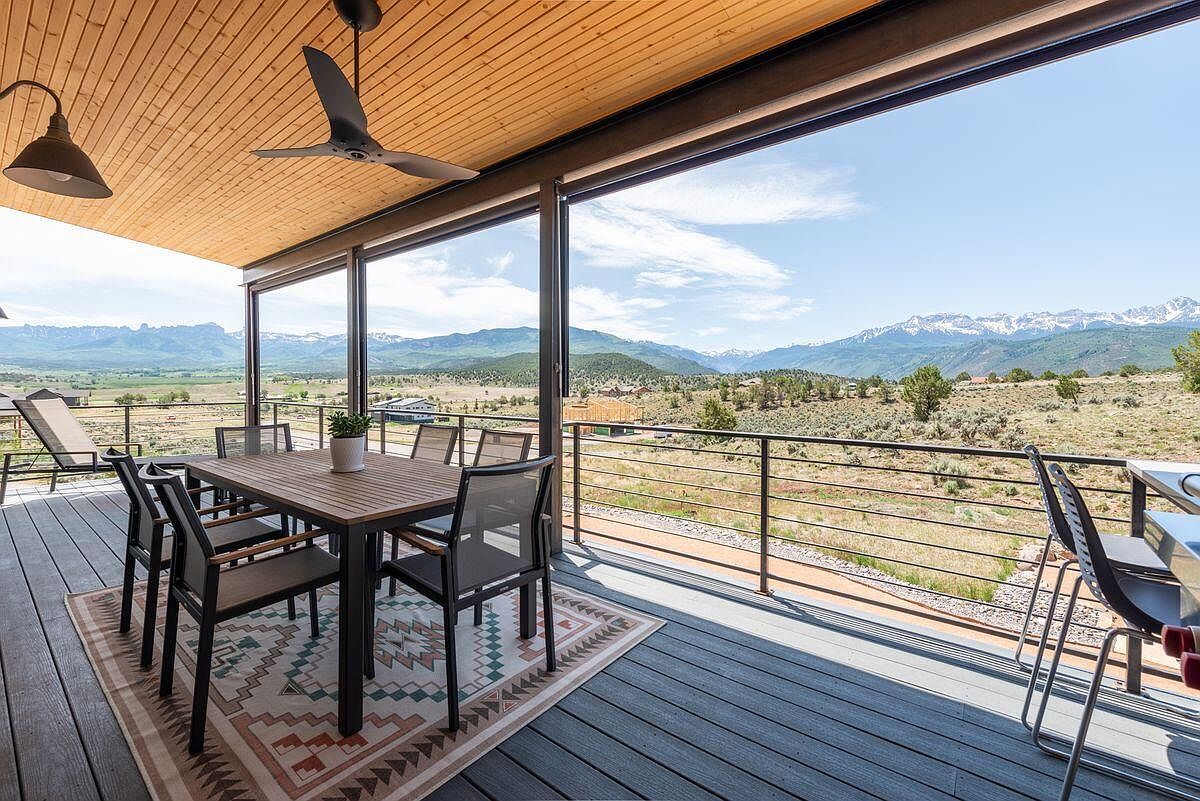This image showcases a stunning outdoor patio or deck area, featuring a dining table set for six, a ceiling fan, and expansive sliding glass doors that frame a breathtaking mountain view. The deck is constructed with modern gray composite decking, complemented by a stylish outdoor rug, creating an inviting space for relaxation and entertainment. The open design seamlessly blends indoor and outdoor living, making it a desirable feature for potential buyers.
