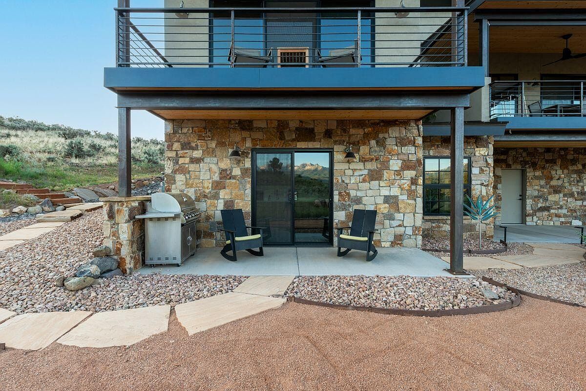This is an exterior shot of a patio area featuring stone accents and modern design elements. The patio includes outdoor seating, a built-in grill, and a sliding glass door leading into the home. The space is surrounded by gravel and stone pathways, creating a visually appealing and functional outdoor living area.