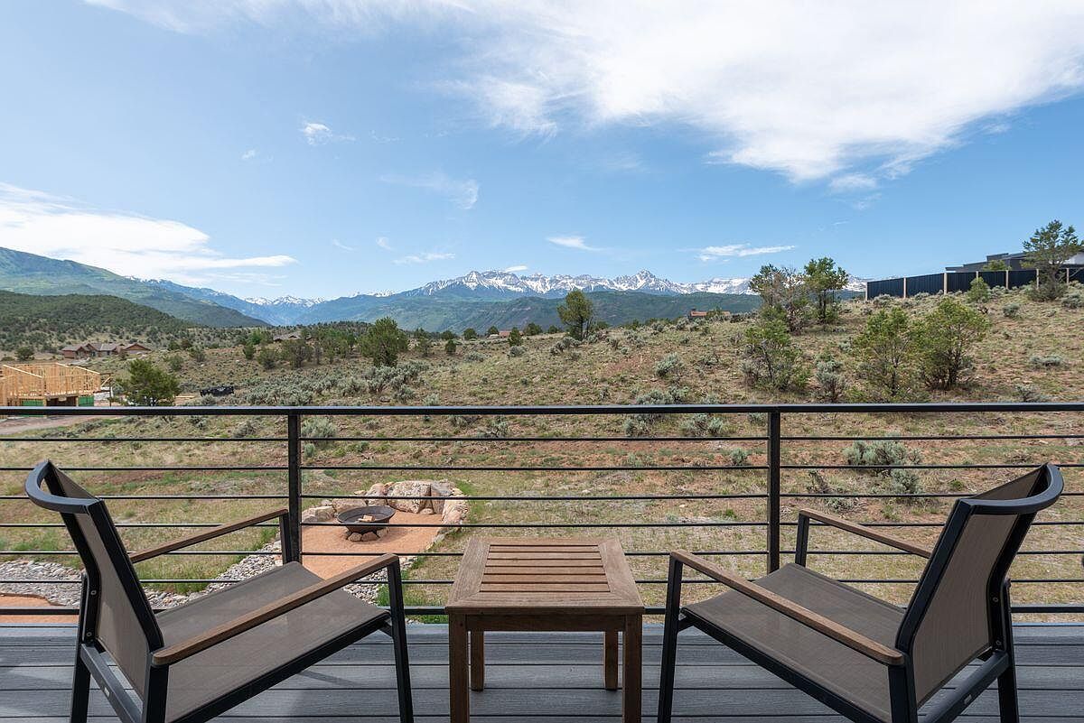 This image showcases a serene outdoor living space, specifically a patio or deck, with a breathtaking mountain view. Two modern chairs and a small wooden table are arranged on the deck, inviting relaxation and enjoyment of the scenery. A fire pit is visible in the yard below, suggesting a cozy gathering spot.
