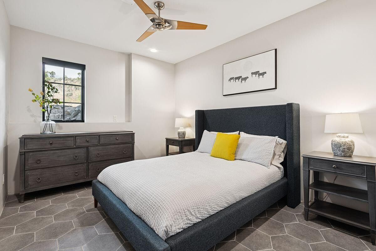 This is a well-lit primary bedroom featuring a large bed with a gray upholstered headboard and a yellow accent pillow. The room includes a dark wood dresser, a matching nightstand with a lamp, and a framed piece of art above the bed. The flooring is a unique gray tile pattern, and a window provides natural light, creating a cozy and inviting atmosphere.
