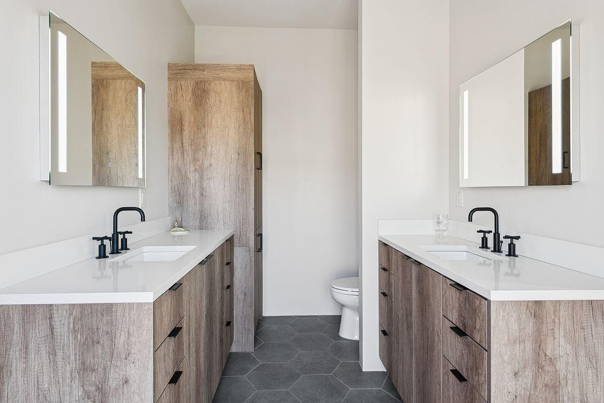 This is a well-lit primary bathroom featuring two separate vanities with wood-grain cabinets, white countertops, and modern black faucets. Each vanity has a rectangular mirror with integrated lighting. The floor is covered in gray hexagonal tiles, and a tall wooden cabinet stands between the vanities, adding storage space.