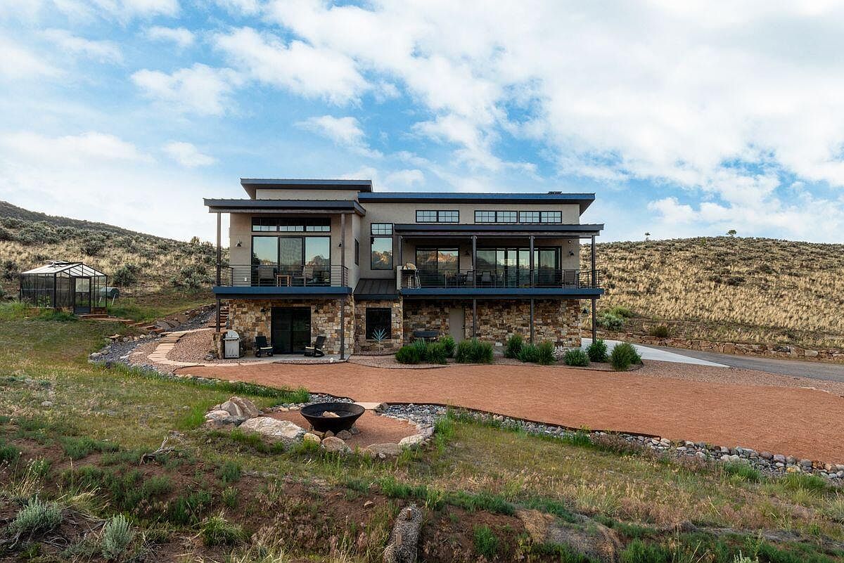 This is a rear view of a modern two-story home with a stone facade on the lower level and stucco on the upper level. The house features balconies with metal railings, large windows, and a flat roof. The landscaping includes a gravel area, a fire pit, and natural vegetation, blending the home into the surrounding hillside.