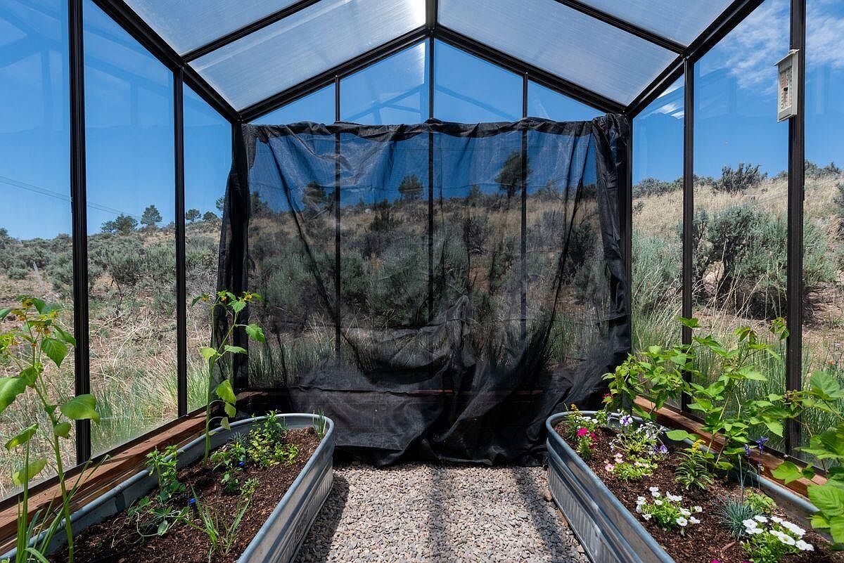 This image showcases the interior of a greenhouse, featuring two galvanized metal planters filled with lush greenery and colorful flowers. The greenhouse structure is made of glass panels with a black frame, allowing ample sunlight to filter through. A black mesh screen is positioned at the back, partially obscuring the view of the natural landscape outside, creating a sense of privacy and controlled environment for plant growth.