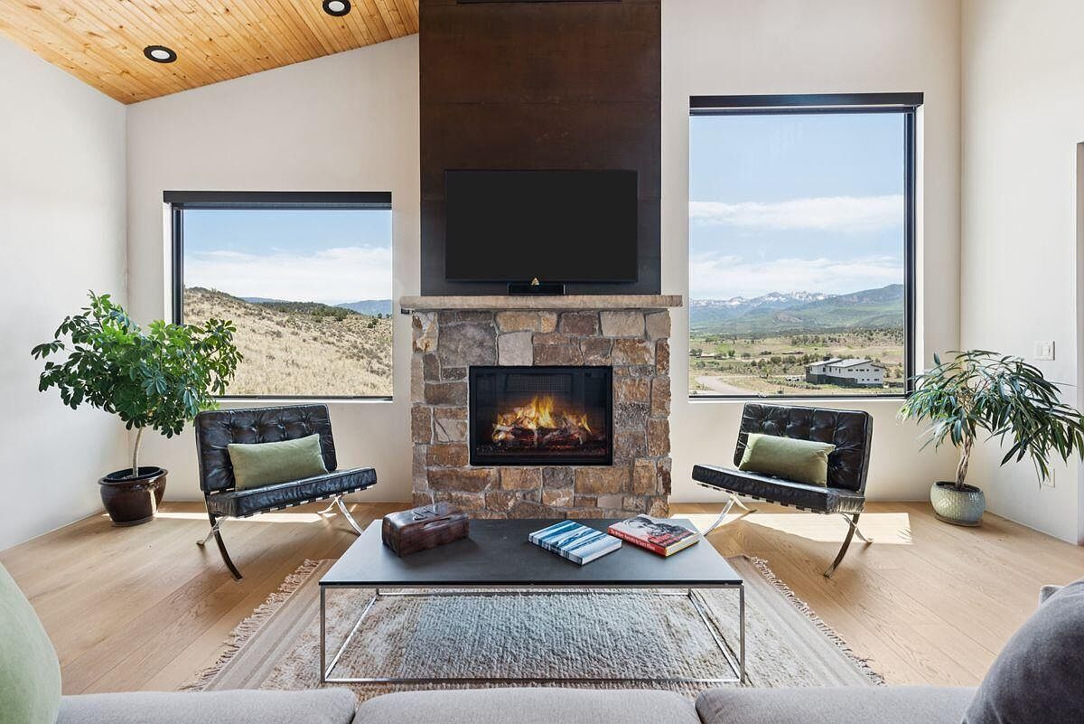 This is an interior shot of a living room featuring a stone fireplace with a mounted television above it. Two modern black leather chairs flank the fireplace, and a coffee table sits in front of them on a patterned rug. Large windows offer views of the landscape outside, and the room has a modern, minimalist aesthetic with light wood floors and a vaulted ceiling.