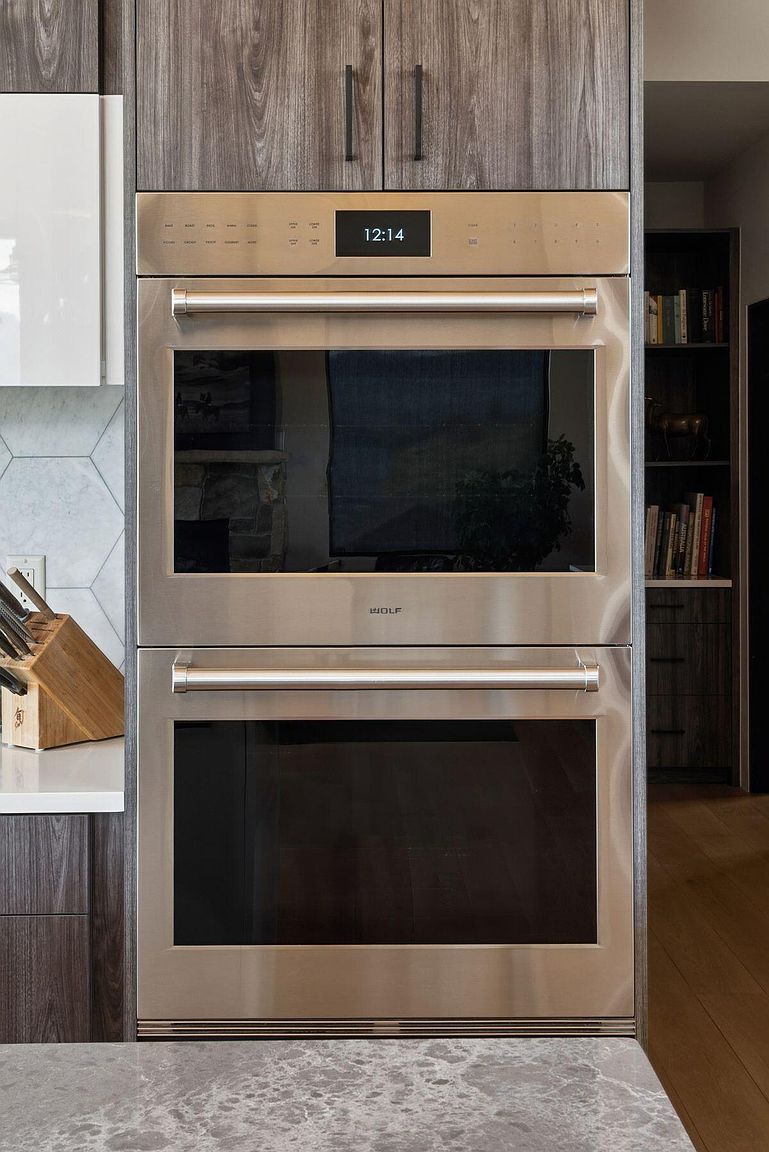The image showcases a built-in double oven, likely a Wolf brand, with a stainless steel finish. The ovens are integrated into wooden cabinetry, and the digital display shows the time. The overall impression is one of high-end kitchen appliances and modern design.