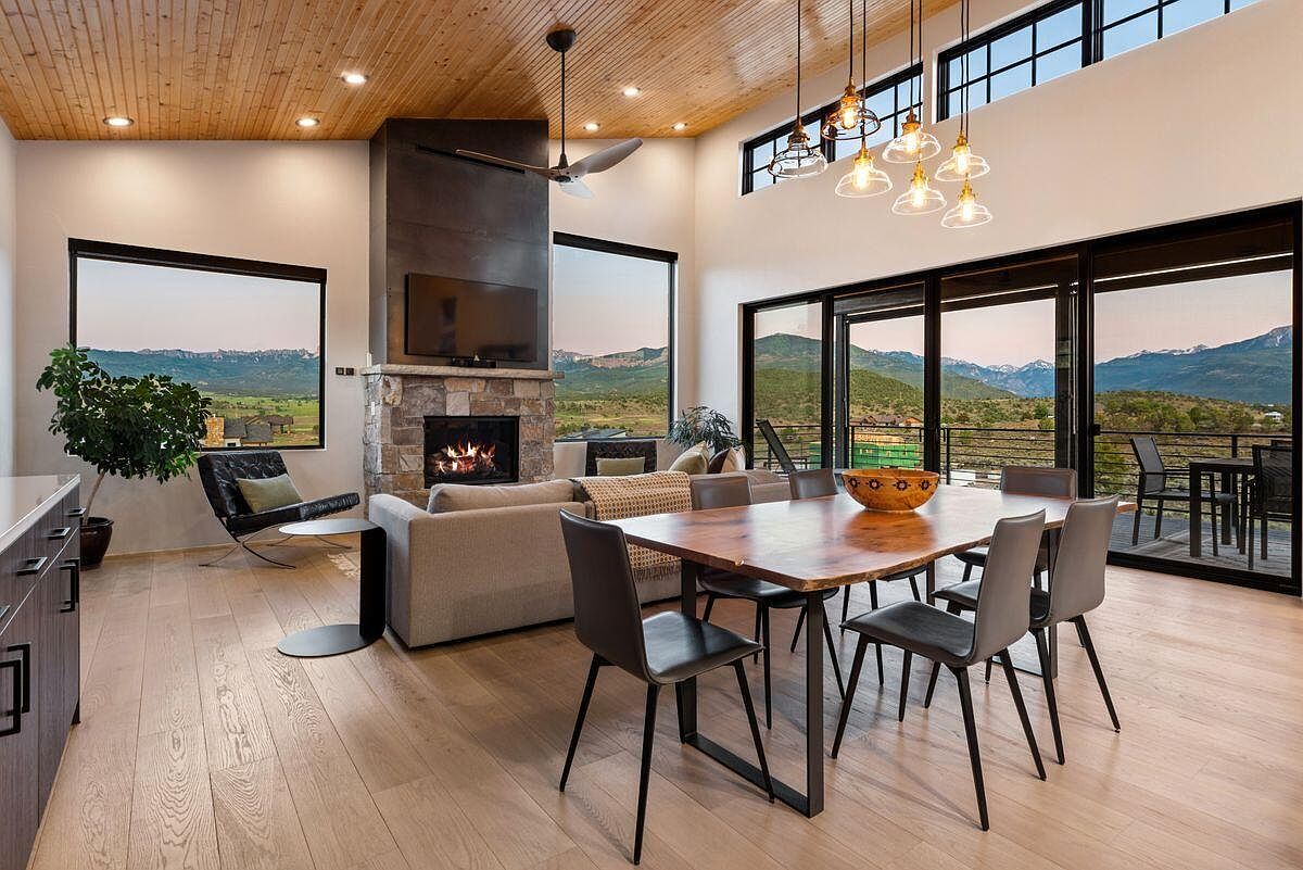 This is an interior shot of a luxurious living and dining area, featuring a stone fireplace with a mounted TV, a comfortable sofa, and a modern dining table with stylish chairs. Large windows and sliding glass doors offer stunning views of the surrounding landscape, creating a seamless connection between indoor and outdoor living. The room is well-lit with a combination of natural light and pendant lighting, enhancing the warm and inviting atmosphere.