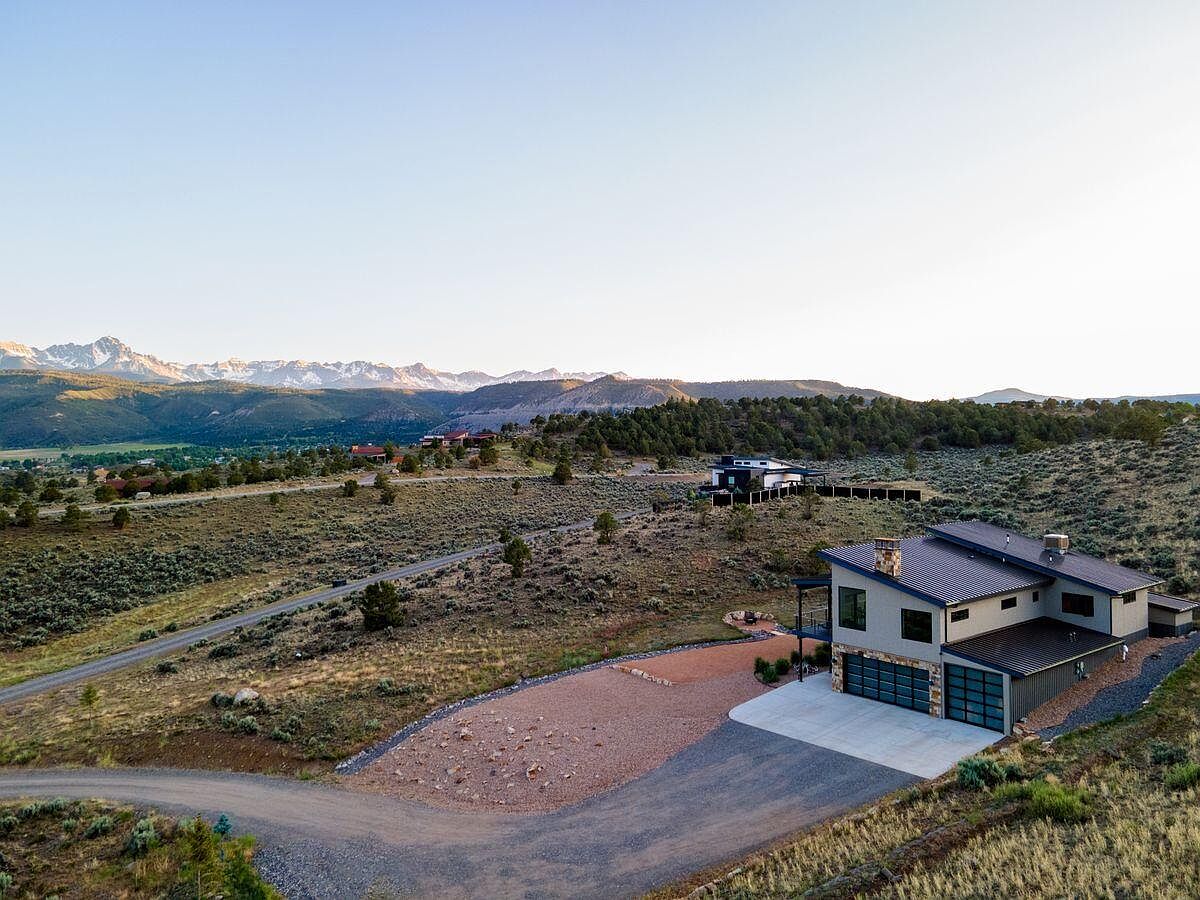 This aerial view showcases a modern home nestled in a scenic landscape with mountains in the background. The property features a spacious driveway, a well-maintained yard, and a contemporary architectural design with a combination of light and dark exterior elements. The surrounding terrain is a mix of natural vegetation and open space, providing a sense of privacy and tranquility.