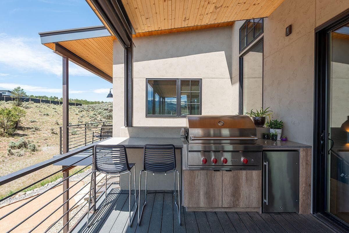 This image showcases a modern outdoor patio or deck area, featuring a built-in grill station with stainless steel appliances and a sleek countertop. Two bar stools are positioned at the counter, offering a space for outdoor dining or relaxation. The deck is surrounded by a railing, providing safety and a clear view of the natural landscape beyond.