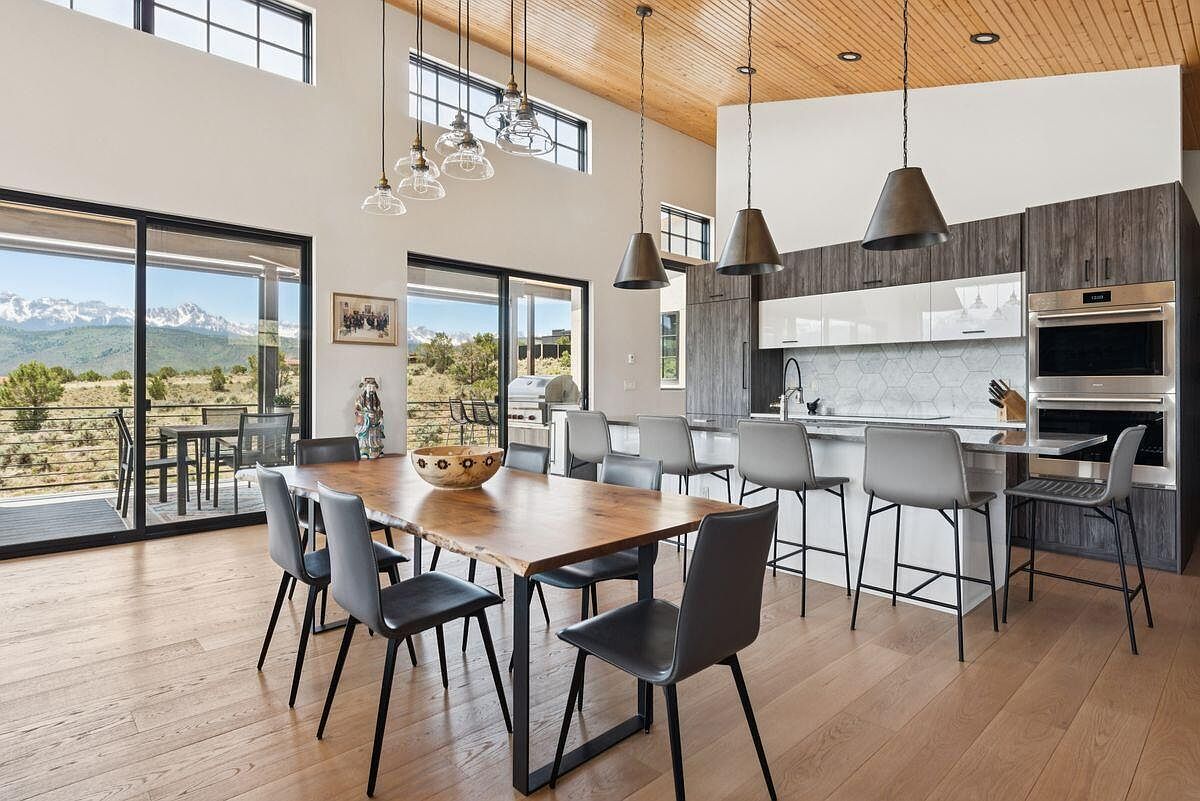 This interior shot showcases a modern dining room seamlessly integrated with the kitchen area. The dining room features a large wooden table with black metal legs, surrounded by gray chairs, and is illuminated by pendant lights. Large sliding glass doors offer stunning views of the landscape, creating an open and airy atmosphere, while the kitchen boasts sleek cabinetry and modern appliances.