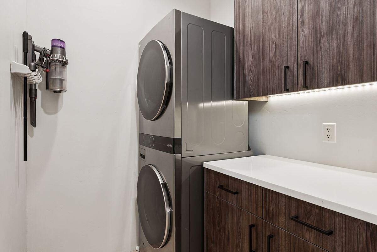 This is a well-organized laundry room featuring a stacked washer and dryer set in a modern gray finish. A cordless vacuum cleaner is mounted on the wall for easy access, and the dark wood cabinets with black hardware provide ample storage space. The white countertop offers a clean workspace, and the under-cabinet lighting adds a touch of elegance.