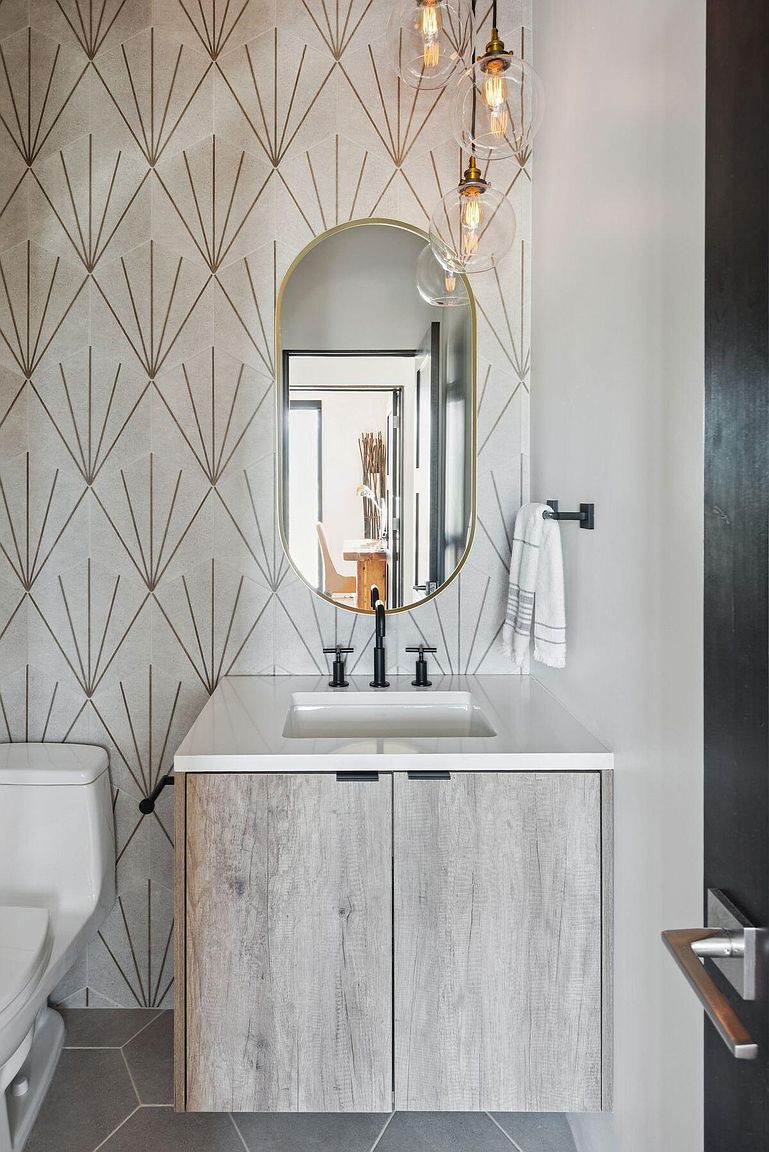 This is a stylish guest bathroom featuring a modern vanity with a light wood-grain finish and a white countertop. An oval mirror with a gold frame hangs above the sink, complemented by a unique geometric tile backsplash. The space is illuminated by pendant lighting, creating a warm and inviting atmosphere.