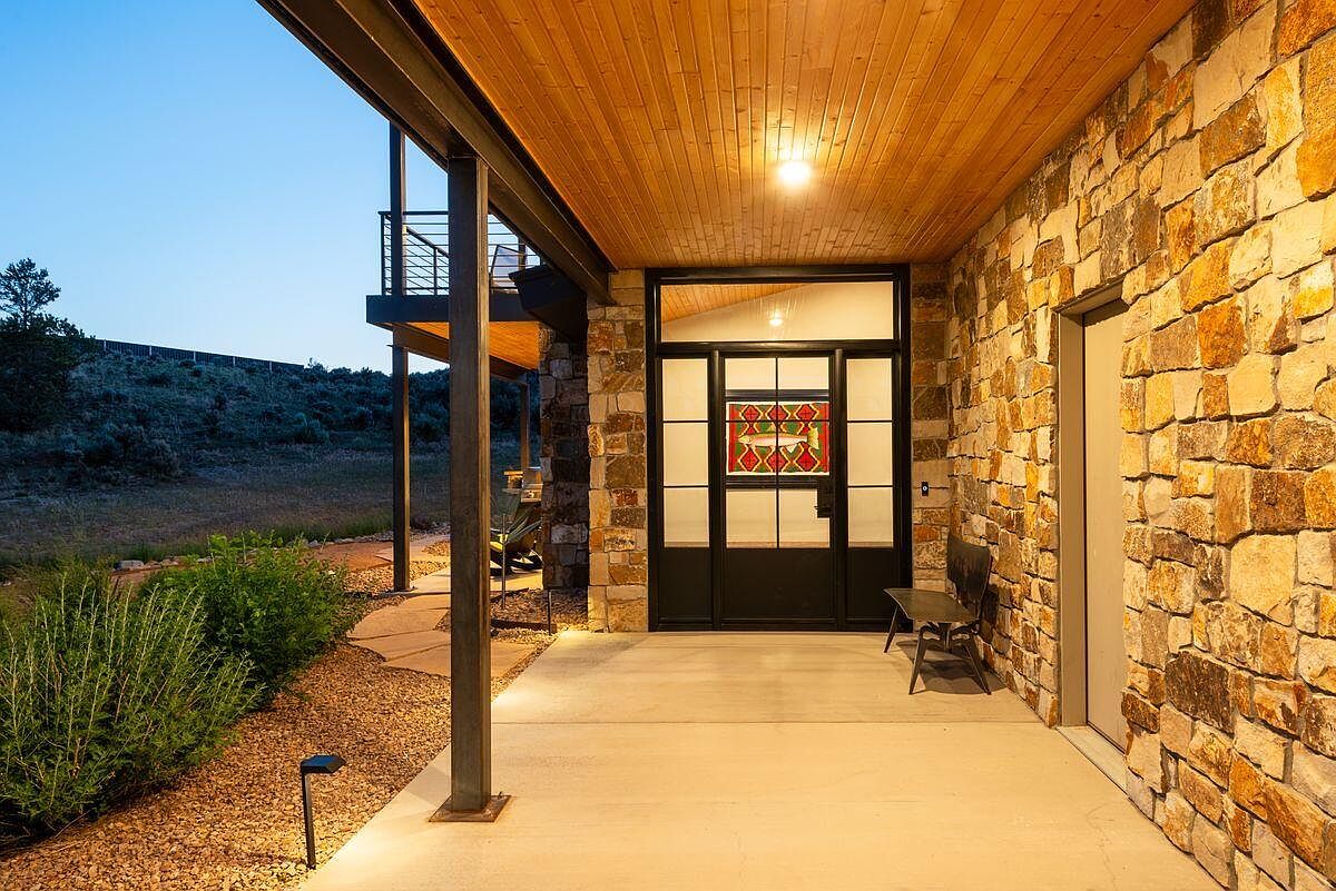 This image showcases the entryway of a home, featuring a stone facade and a wooden ceiling. A black-framed door with decorative glass is the focal point, flanked by a stone wall and a glimpse of the surrounding landscape. The entryway is well-lit and appears inviting, suggesting a blend of rustic charm and modern design.