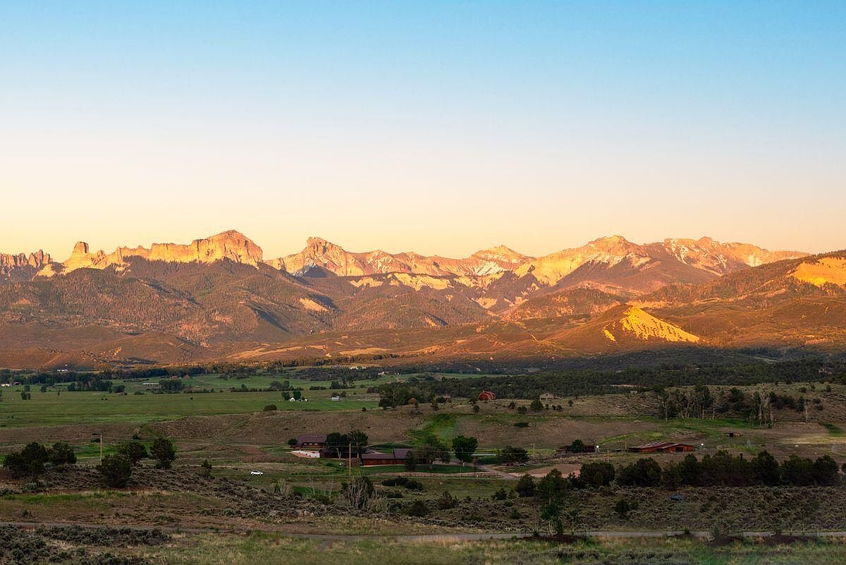 This aerial view showcases a sprawling property nestled in a valley, with a backdrop of majestic mountains bathed in the warm light of sunrise or sunset. The property features several buildings, including what appears to be a main house and outbuildings, surrounded by a mix of green fields and natural terrain. The overall impression is one of serene privacy and connection to nature, highlighting the expansive land and stunning mountain views.
