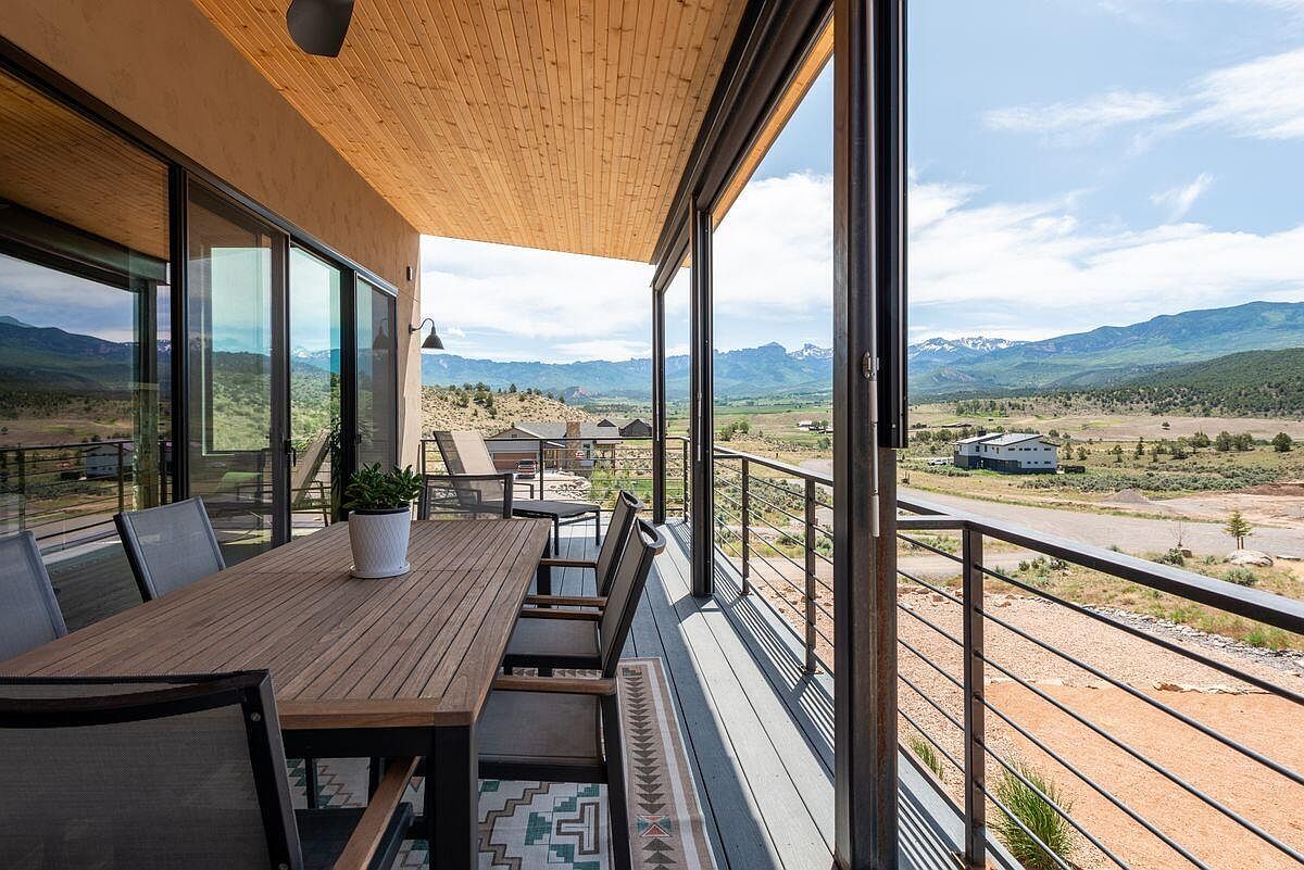 This image showcases a spacious outdoor patio or balcony area, featuring a wooden dining table with chairs, a patterned rug, and a metal railing. The space offers stunning views of the surrounding landscape, including mountains and open fields. The design incorporates modern elements with natural materials, creating an inviting and stylish outdoor living area.