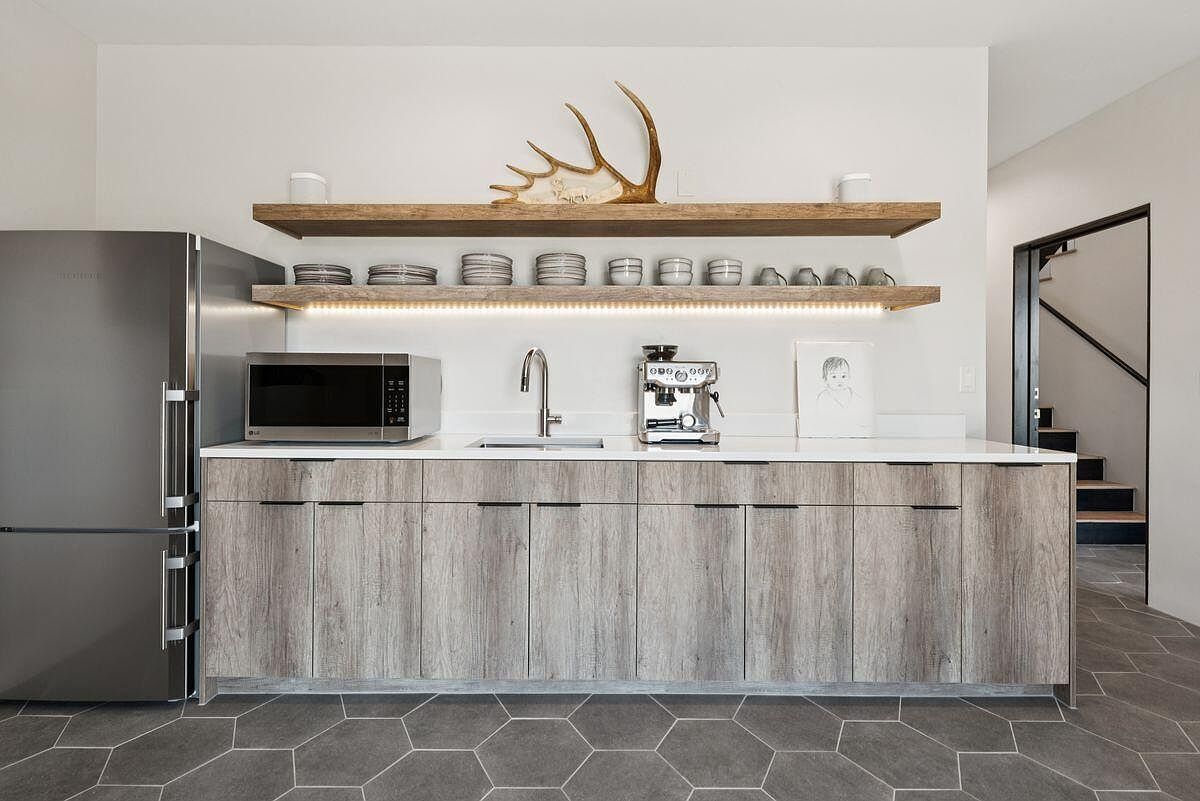 This interior shot showcases a modern kitchen area with a stainless steel refrigerator, microwave, and espresso machine on a white countertop. The cabinets below feature a wood-grain finish with sleek, minimalist hardware. Above the counter, two wooden shelves display decorative items, including plates, cups, and antlers, with under-cabinet lighting adding a warm glow.