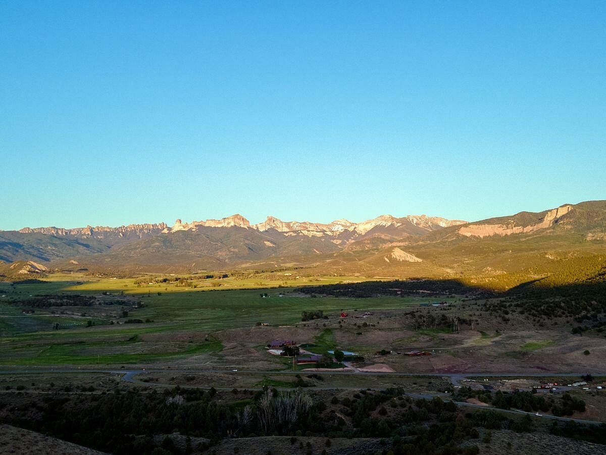 This aerial view showcases a vast landscape with a mountain range in the background, a valley with green fields, and a residential area with houses and roads in the foreground. The scene is bathed in warm sunlight, creating a serene and inviting atmosphere. The perspective gives a sense of the property's location within a larger, picturesque setting.