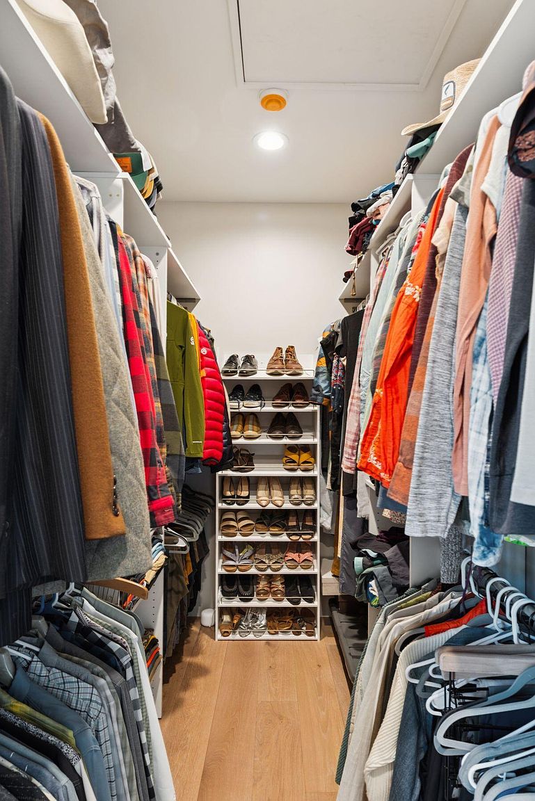 This is a well-organized walk-in closet featuring custom shelving and hanging rods. The closet is filled with a variety of clothing items and shoes, neatly arranged. The light wood flooring and white shelving create a clean and bright space, maximizing storage and accessibility.