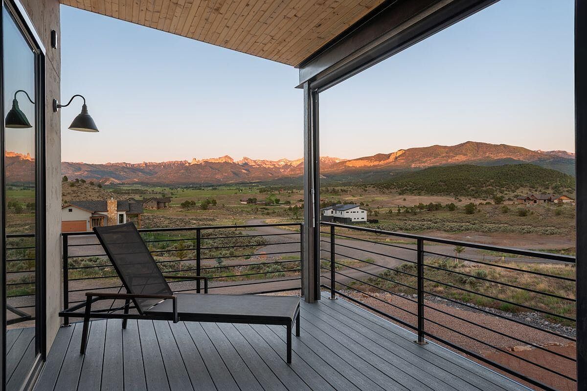 This image showcases a modern balcony with a sleek lounge chair, offering stunning views of a mountain range and valley at sunset. The balcony features a contemporary railing system and a well-maintained deck surface, creating an inviting outdoor living space. The open design seamlessly blends indoor and outdoor living, highlighting the property's connection to its scenic surroundings.