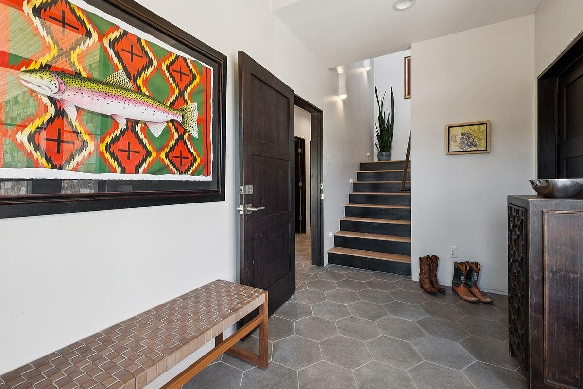 This interior shot showcases a stylish entryway with a striking piece of art featuring a fish, complemented by a woven bench. The hallway leads to a staircase with dark treads and light risers, adding a modern touch. The flooring is a gray hexagon tile, and the overall aesthetic is sophisticated and inviting.