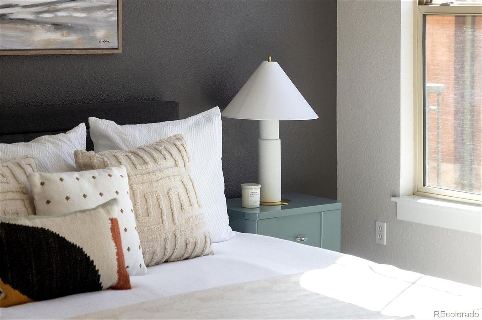 This close-up shot captures a cozy and stylish bedroom corner, featuring a bed layered with textured, neutral-toned throw pillows and a crisp white duvet. A modern, minimalist white table lamp sits atop a sleek, sage-green nightstand, complemented by a small candle and a framed piece of art on the dark accent wall. The composition is intimate and inviting, with soft natural light streaming in from the adjacent window, highlighting the sophisticated color palette and layered textures.