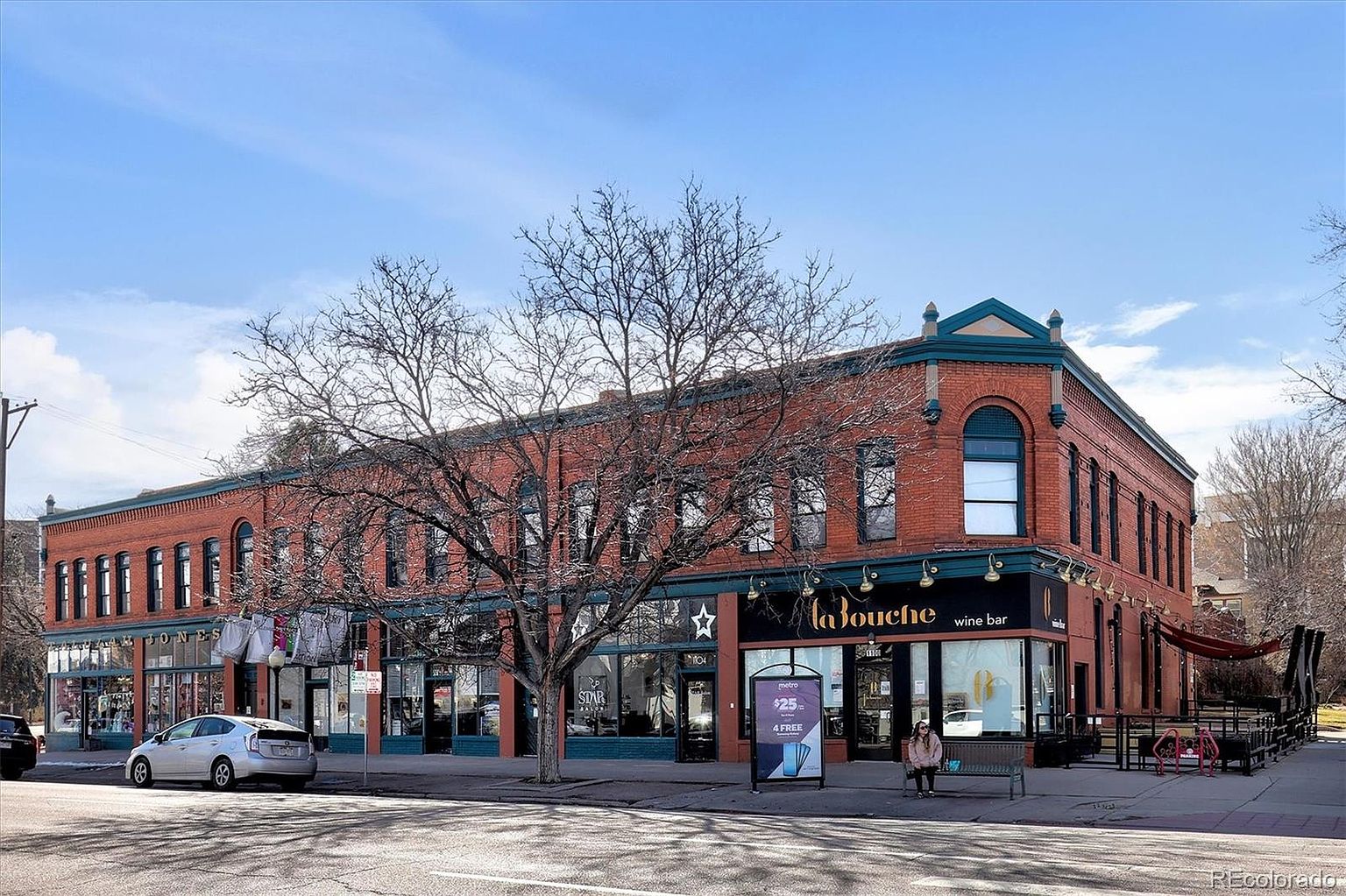 This image captures a prominent corner commercial building constructed of classic red brick, featuring large arched windows on the second floor and storefronts on the ground level. The building houses multiple businesses, including 'La Bouche' wine bar, and is situated on a sunny street corner with a mature tree in the foreground. The perspective is a wide-angle street-level shot that emphasizes the building's historic architectural character and its prominent location in the neighborhood.