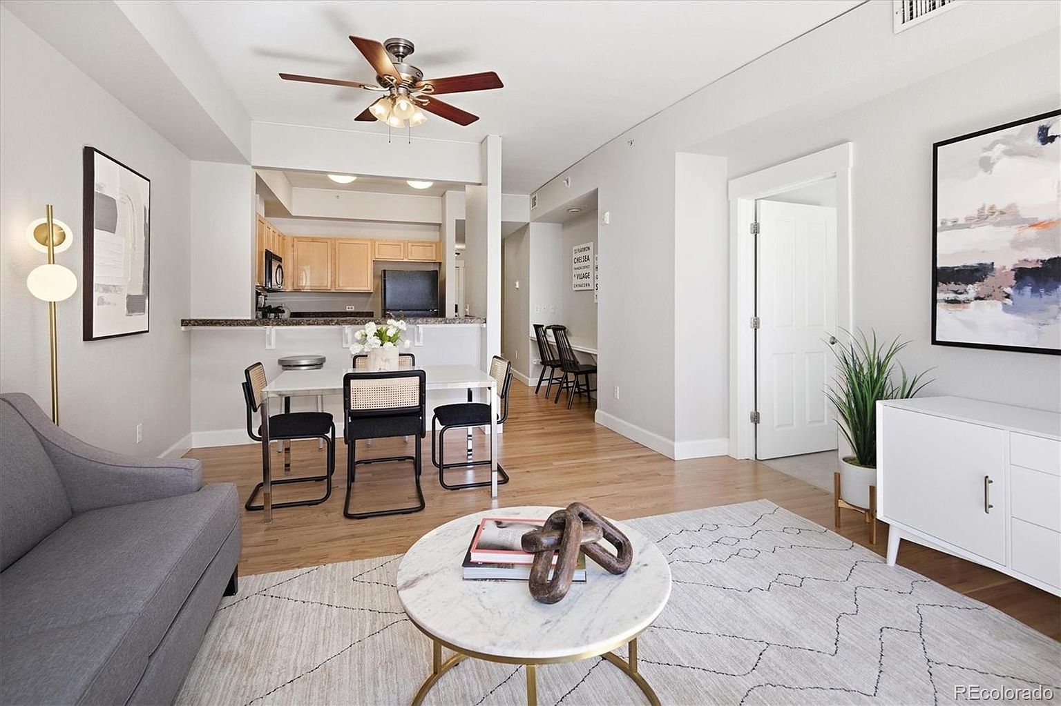 This open-concept living and dining area features a contemporary aesthetic with light wood flooring and neutral walls. The space includes a comfortable gray sofa, a marble-topped coffee table with a sculptural centerpiece, and a dining set with modern cane-back chairs, all illuminated by a ceiling fan and stylish floor lamp. The perspective captures the flow from the living area into the kitchen and a small office nook, creating a bright and inviting atmosphere.