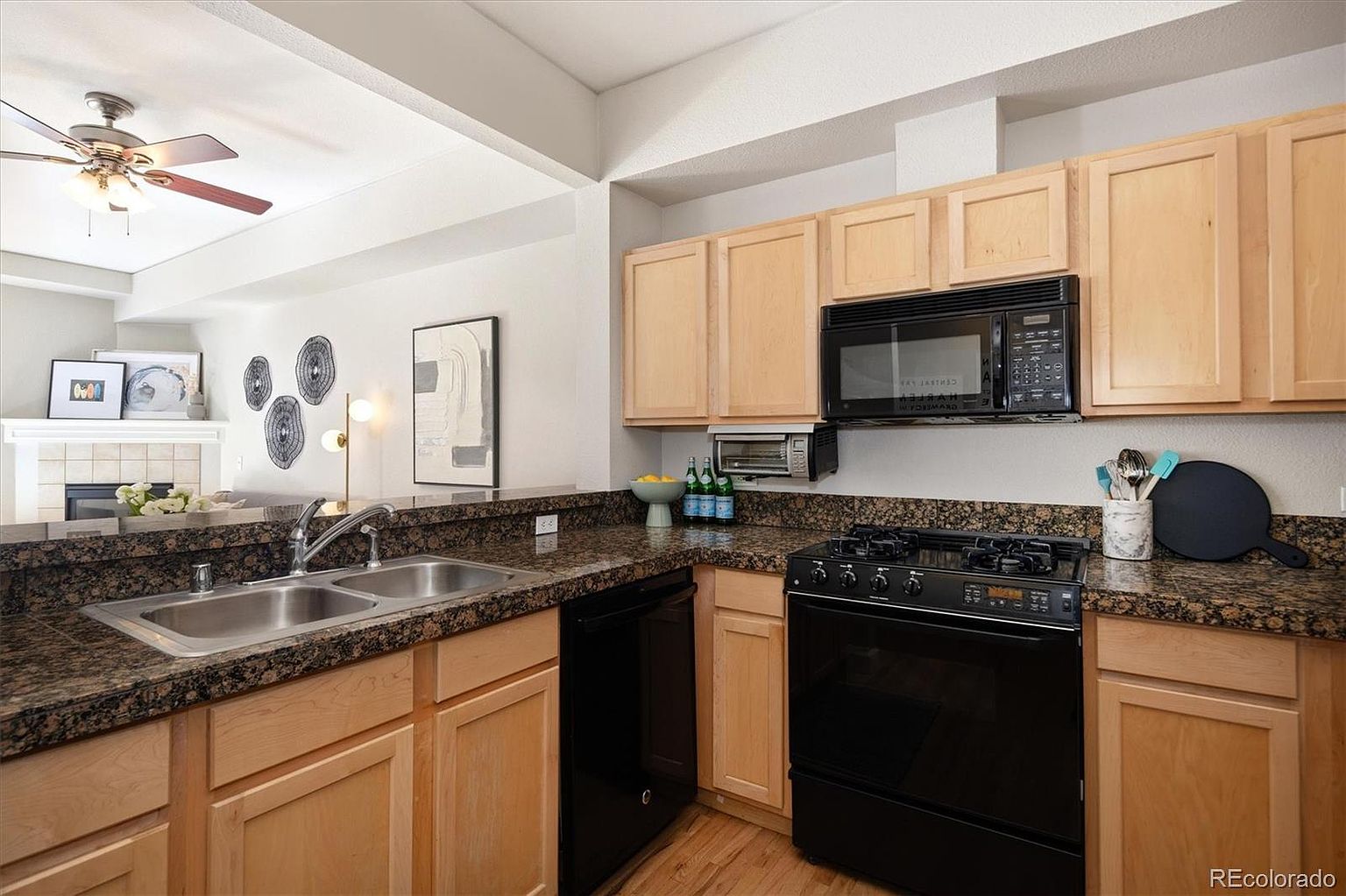 This kitchen features light wood cabinetry paired with dark granite countertops and black appliances, creating a functional and classic aesthetic. The space includes a double-basin sink, a gas range, and an open-concept layout that looks out toward the living area. The perspective is captured from a standing position, highlighting the efficient workflow and the connection between the kitchen and the adjacent living space.