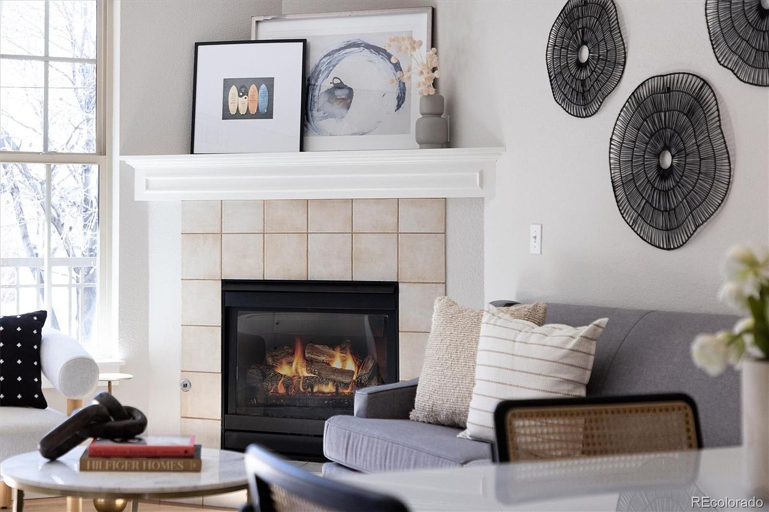 This cozy living room features a central fireplace with a light-colored tile surround and a classic white mantel, topped with curated art and decor. A comfortable grey sofa with textured throw pillows sits adjacent to the fireplace, while a round coffee table with a book adds a touch of sophistication. The space is brightened by natural light from a large window, creating a warm and inviting atmosphere perfect for relaxation.