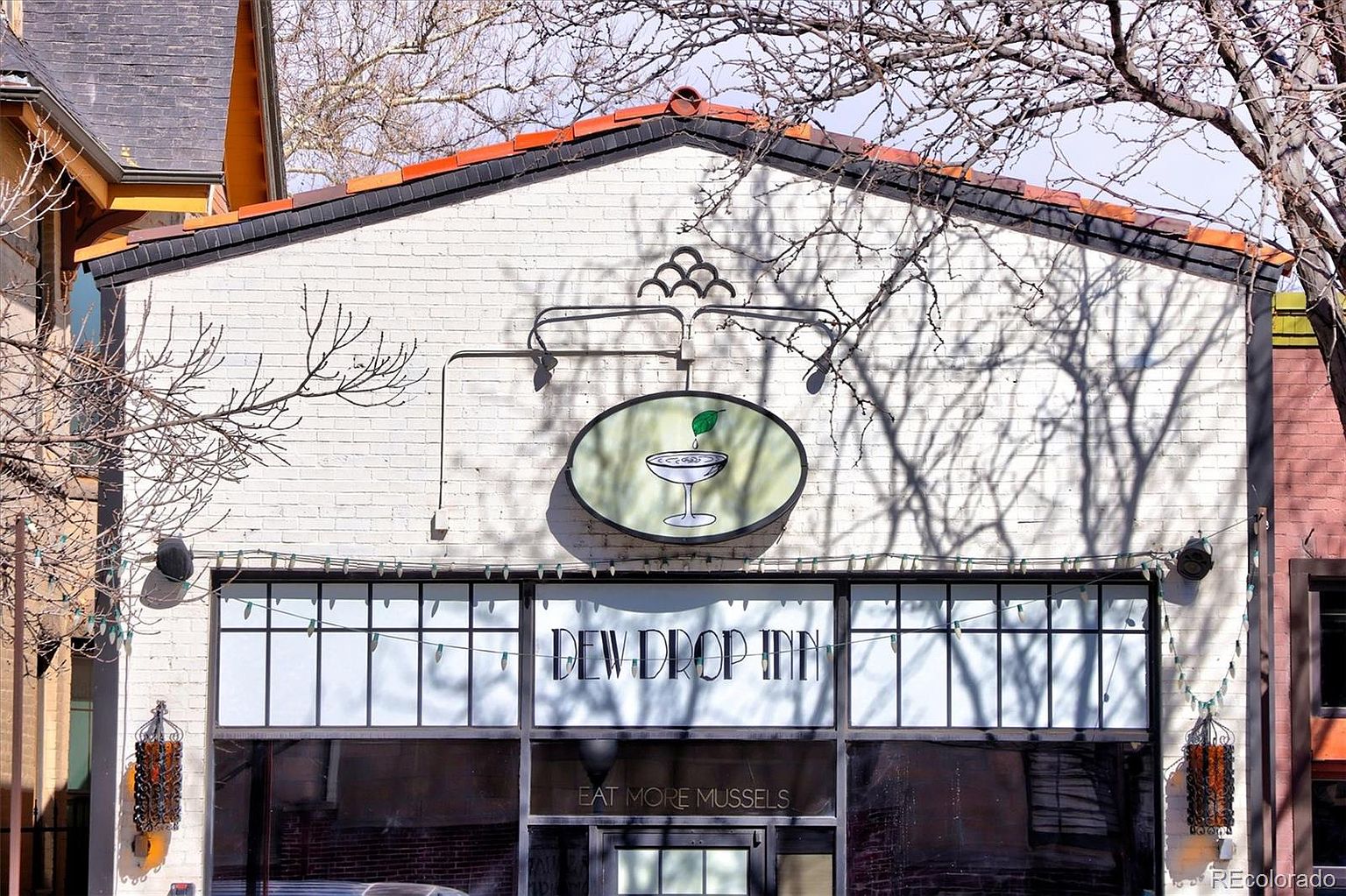 This image captures the front facade of the 'Dew Drop Inn,' a charming commercial building featuring a white-painted brick exterior and a distinctive, arched roofline. A prominent oval sign depicting a martini glass with a leaf hangs above the large, multi-paned windows, which are adorned with string lights and the phrase 'EAT MORE MUSSELS' etched on the glass. The perspective is a straight-on, eye-level shot that highlights the building's unique character and inviting storefront appeal.