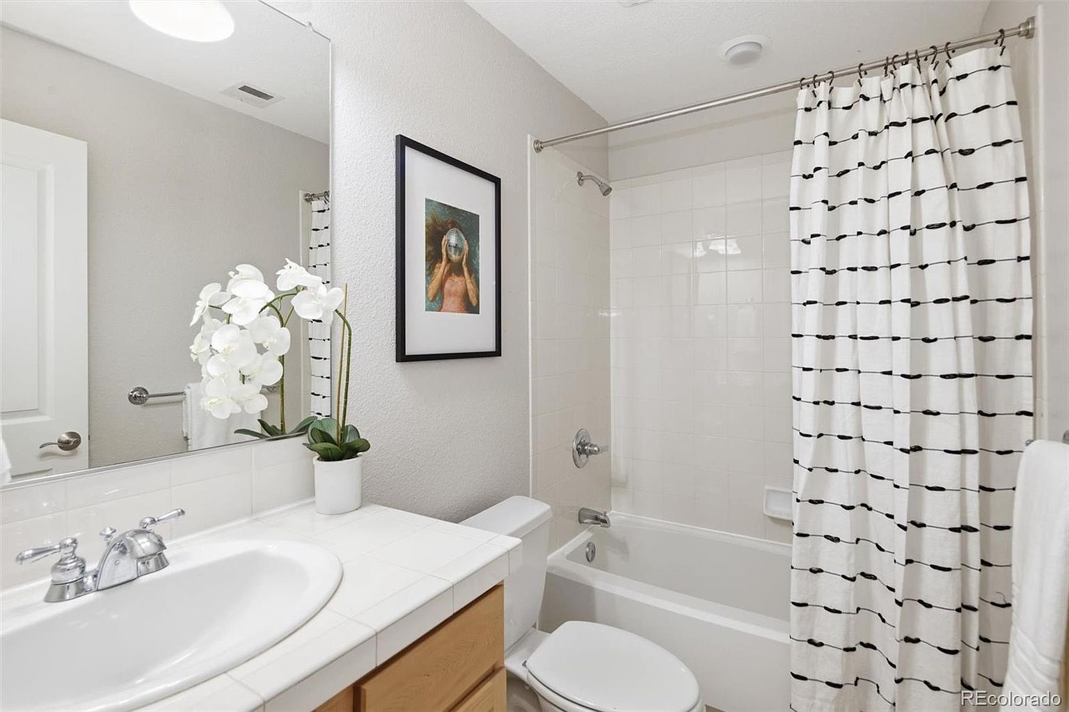 This clean and functional bathroom features a white tiled vanity countertop, a single sink with chrome fixtures, and a framed mirror above. To the right, a shower-tub combination is enclosed by a modern, patterned shower curtain, creating a bright and tidy aesthetic. The space is accented with a small potted orchid and a framed piece of wall art, offering a welcoming and well-maintained appearance.