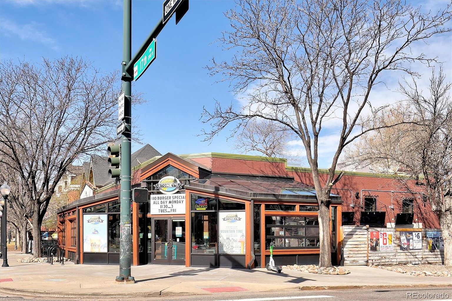 This image captures the front corner view of Stoney's, a commercial establishment with a brick facade and large glass windows. The building features a prominent entrance with signage, a street corner location with a traffic light pole, and a sidewalk area with bike racks and a scooter. The scene is set on a clear, sunny day with bare trees, suggesting a vibrant urban neighborhood setting.