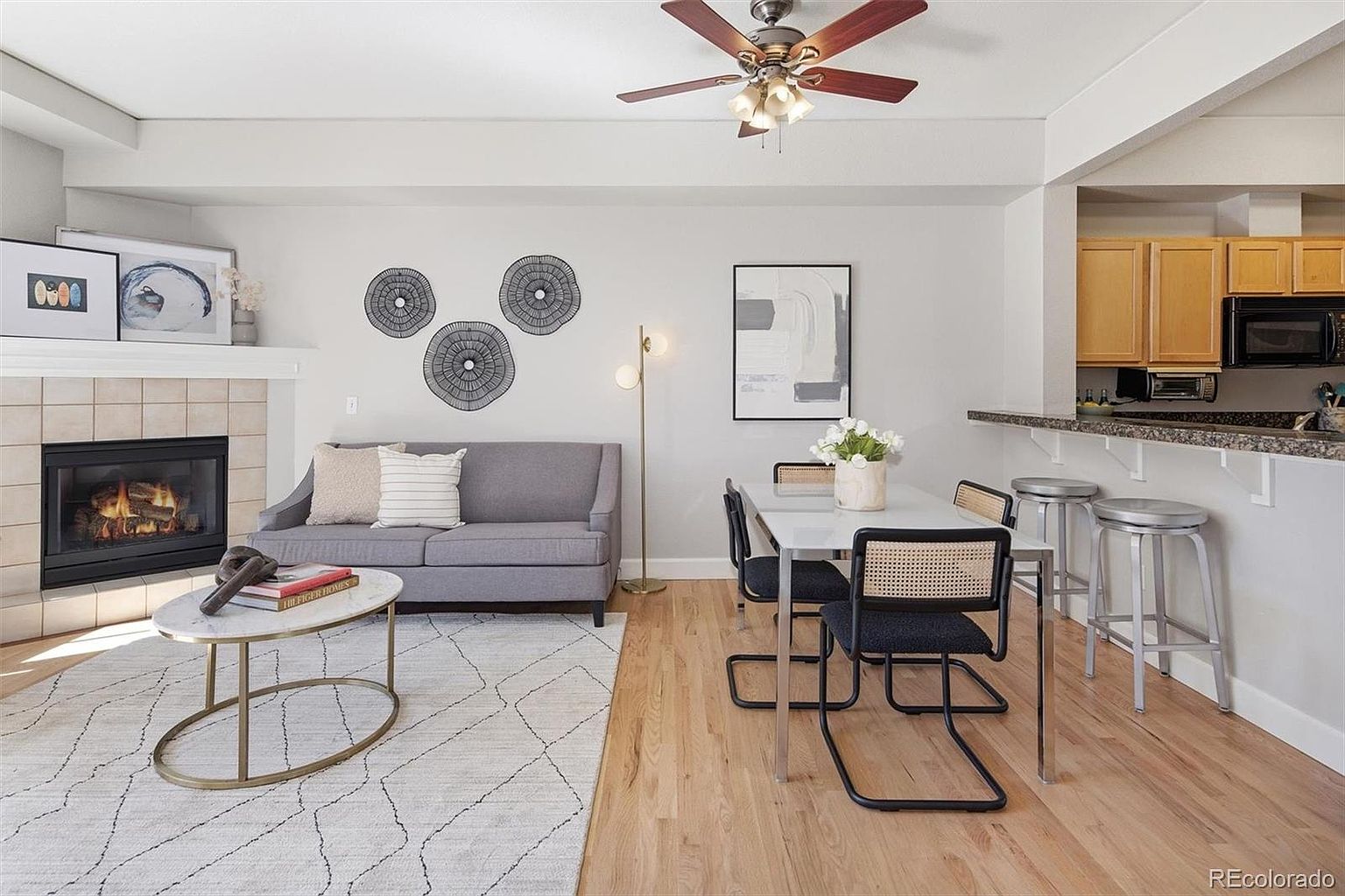 This open-concept living and dining area features a cozy gray sofa positioned in front of a fireplace with a tiled surround. The space is bright and airy, showcasing light hardwood floors, a modern dining set with cane-back chairs, and a breakfast bar that connects to the kitchen. The neutral color palette and contemporary decor create a welcoming and functional living environment.