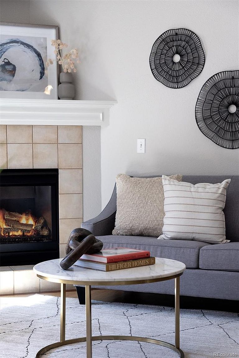 This cozy living room features a modern gray sofa adorned with textured and striped throw pillows, positioned next to a fireplace with a tiled surround. A round marble-topped coffee table with a gold base sits in the foreground, holding a decorative chain and a book, while the wall is accented with two circular black wire art pieces. The scene is captured from a slightly elevated, eye-level perspective, emphasizing a warm, inviting, and contemporary aesthetic.