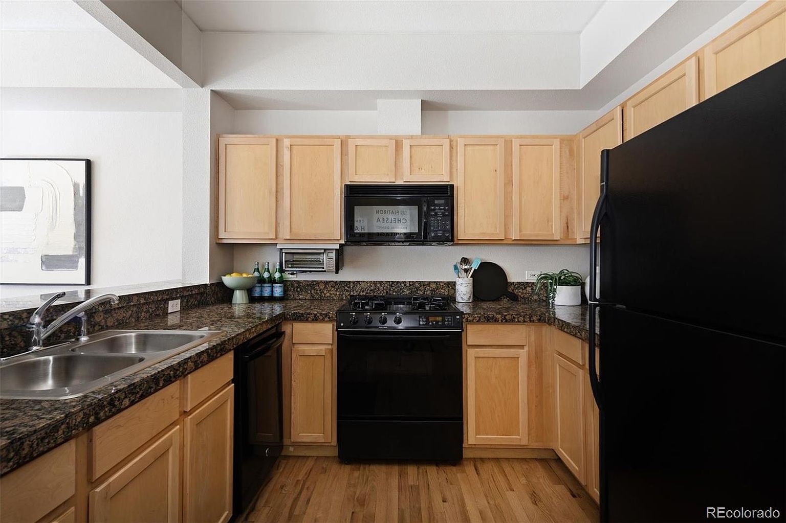 This functional kitchen features light wood cabinetry paired with dark granite countertops and black appliances, creating a high-contrast aesthetic. The U-shaped layout includes a double-basin stainless steel sink and a built-in microwave, offering a practical workspace. The perspective is a straight-on, eye-level shot that highlights the efficient use of space and the warm, natural wood flooring.