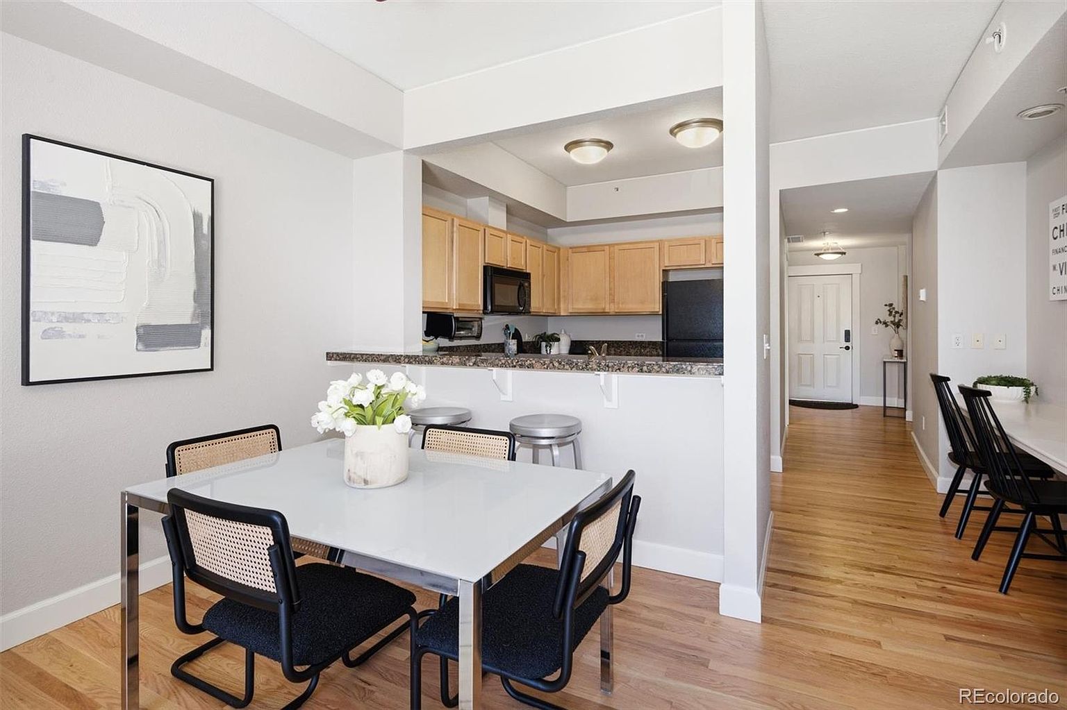 This bright and open-concept dining area features a modern glass-top table with black cane-back chairs, set against a backdrop of light hardwood flooring. The space seamlessly transitions into a functional kitchen with light wood cabinetry and a granite breakfast bar, while a hallway leads toward the entryway. The overall aesthetic is clean, contemporary, and inviting, making it an ideal space for both casual dining and entertaining.