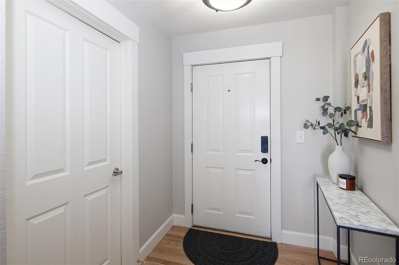 This bright and welcoming entryway features a clean, modern aesthetic with light gray walls and warm wood flooring. The space includes a white front door with a smart lock, a sleek marble-topped console table adorned with a vase of eucalyptus and a candle, and a piece of abstract wall art. A dark, semi-circular rug sits at the base of the door, creating a functional and stylish transition area.