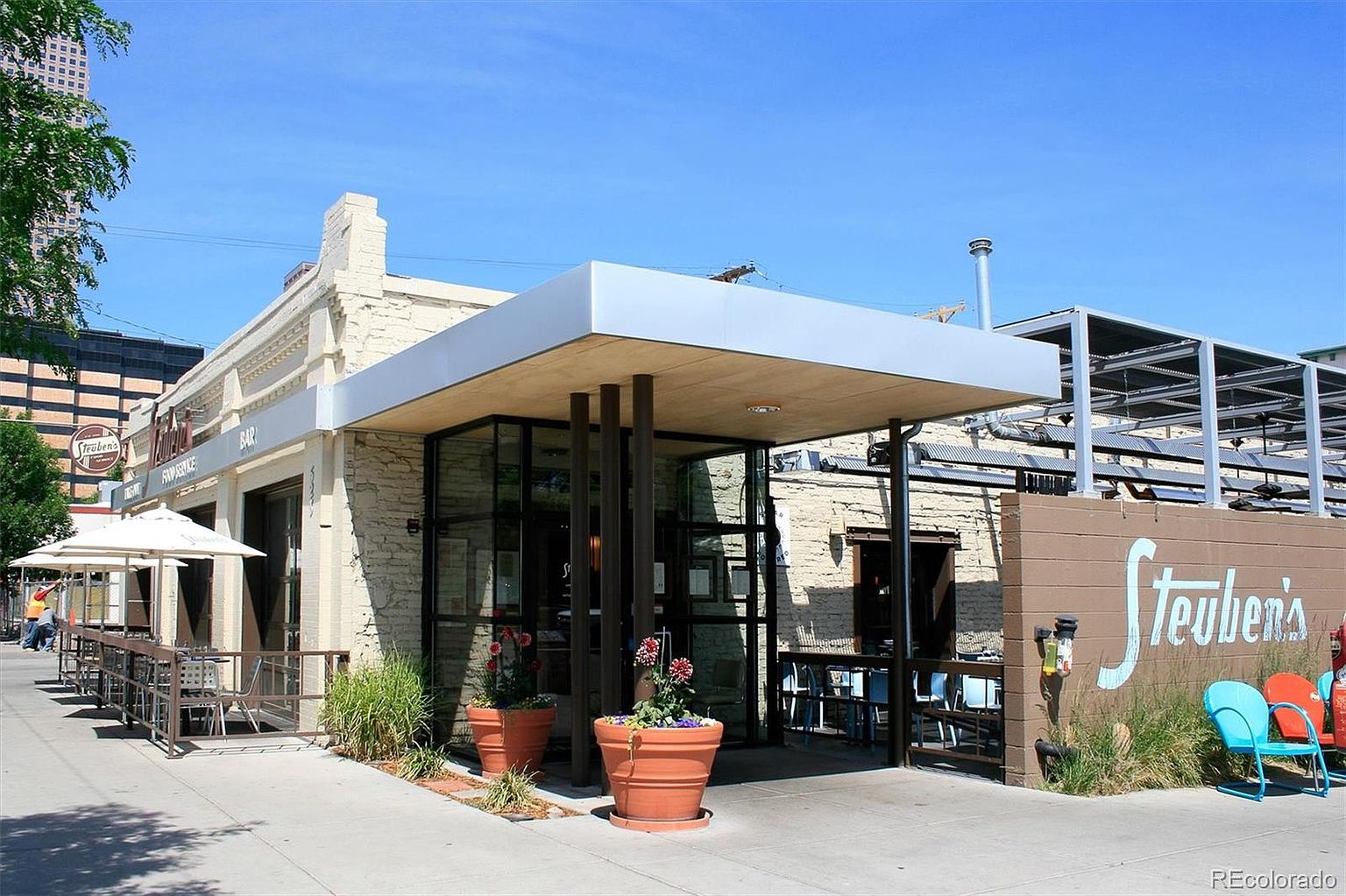 This image captures the front exterior of Steuben's restaurant, showcasing a blend of historic brick architecture and a modern, flat-roofed entryway canopy. The scene features an inviting outdoor patio area with white umbrellas, metal seating, and vibrant potted flowers, set against a clear blue sky. The perspective is a wide-angle street-level shot that highlights the establishment's welcoming curb appeal and urban character.