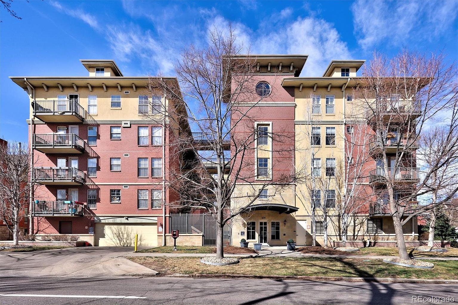 This image captures the front facade of a multi-story residential apartment building, showcasing a blend of red brick and tan stucco architectural elements. The structure features symmetrical balconies, a central tower with a circular window, and a welcoming ground-level entryway, all set against a clear blue sky. The perspective is a straight-on, eye-level shot that highlights the building's urban design and well-maintained landscaping.