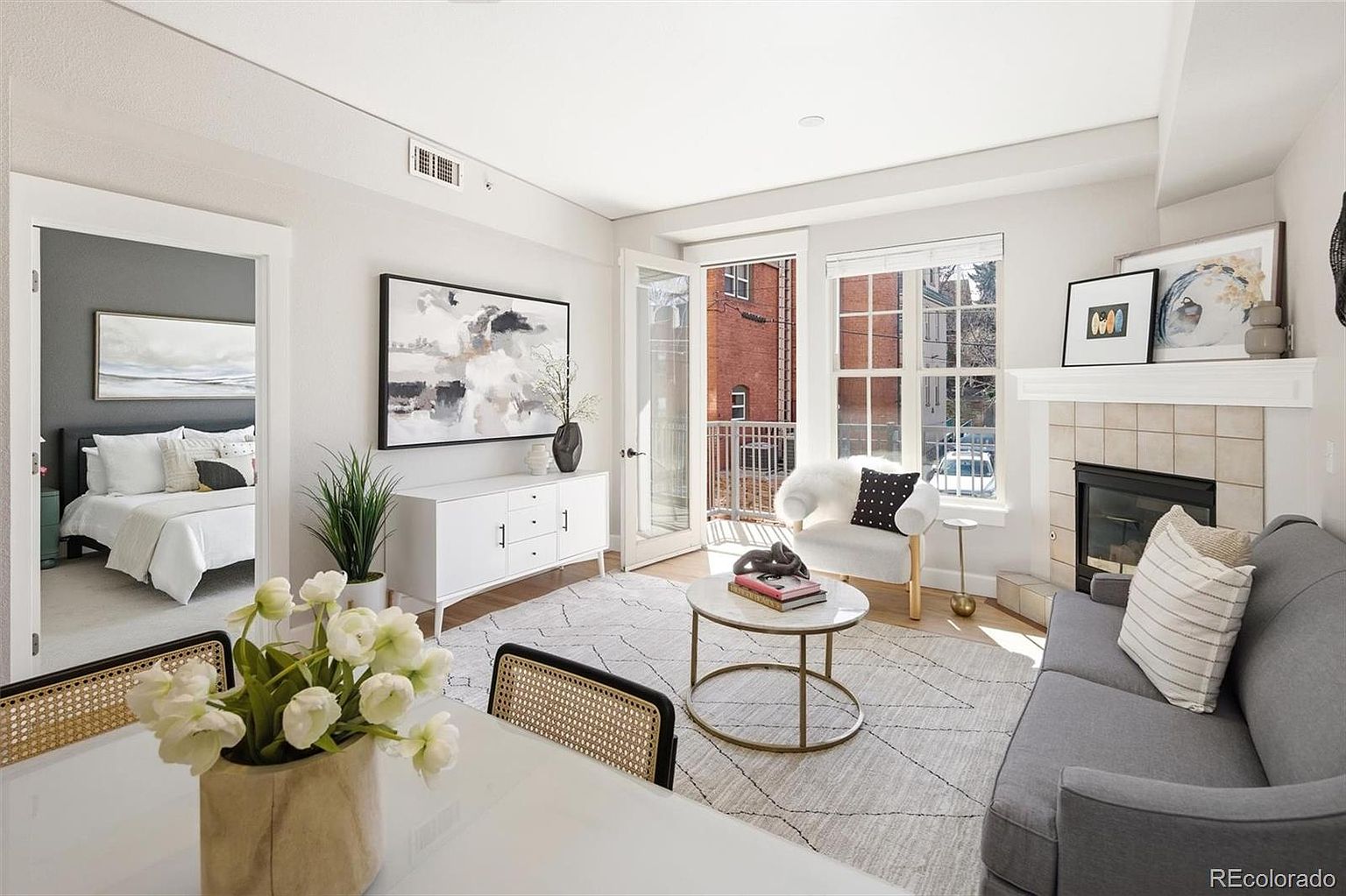 This bright and airy living room features a contemporary design with a neutral color palette, highlighted by a cozy gray sofa, a marble-topped coffee table, and a plush white accent chair. A fireplace with a tiled surround serves as a focal point, while a glass door leads to a small balcony, inviting natural light into the space. The room seamlessly connects to an adjacent bedroom, creating an open and inviting atmosphere perfect for modern living.