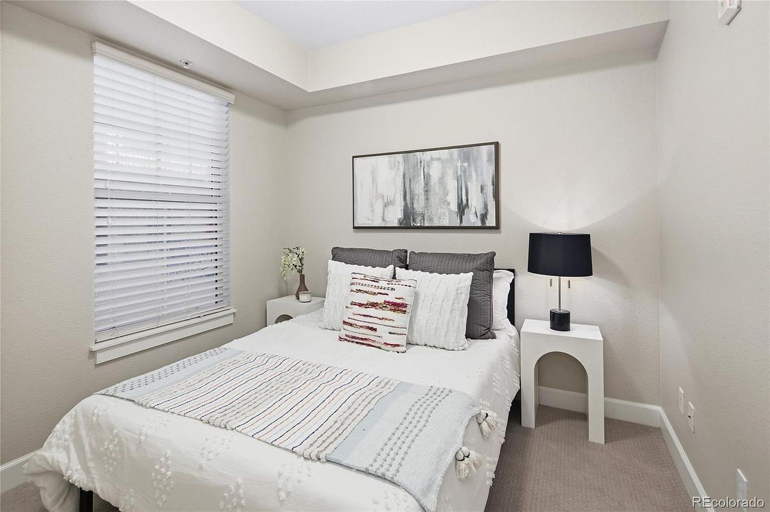 This cozy guest bedroom features a neutral color palette with a queen-sized bed adorned with layered white and grey bedding. A minimalist white nightstand with a modern black lamp sits beside the bed, while a window with white blinds provides natural light. The room is accented by a piece of abstract wall art, creating a clean, inviting, and contemporary atmosphere.