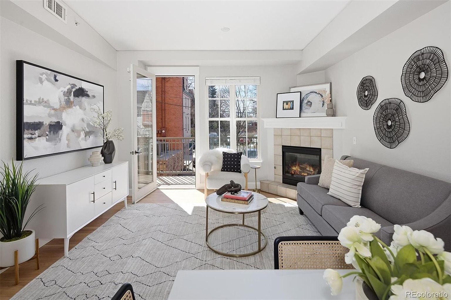 This bright and inviting living room features a contemporary design with a neutral color palette, highlighted by a cozy gray sofa and a white sideboard. A fireplace with a tiled surround serves as a focal point, while a glass door opens to a small balcony, allowing for natural light to fill the space. The room is accented with modern wall art and circular wall decor, creating a sophisticated and comfortable atmosphere.