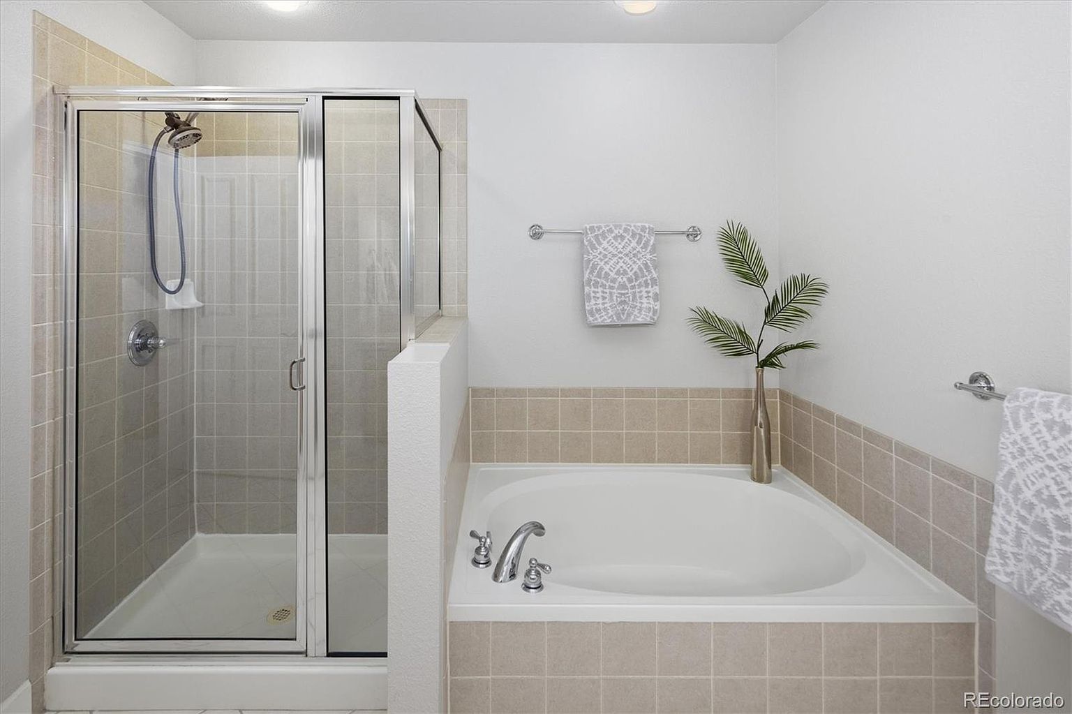 This primary bathroom features a clean, neutral design with a spacious soaking tub and a separate glass-enclosed shower. The area is accented with light beige square tiles that wrap around the tub and shower base, creating a cohesive and calming aesthetic. A decorative towel and a simple vase with greenery add a touch of elegance to the bright, well-lit space.